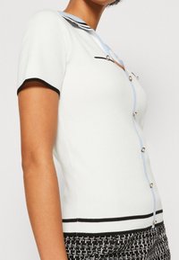Chemise blanche à manches courtes avec un col bleu clair, des accents noirs et des boutons en métal. Texture tricotée avec une coupe ajustée.