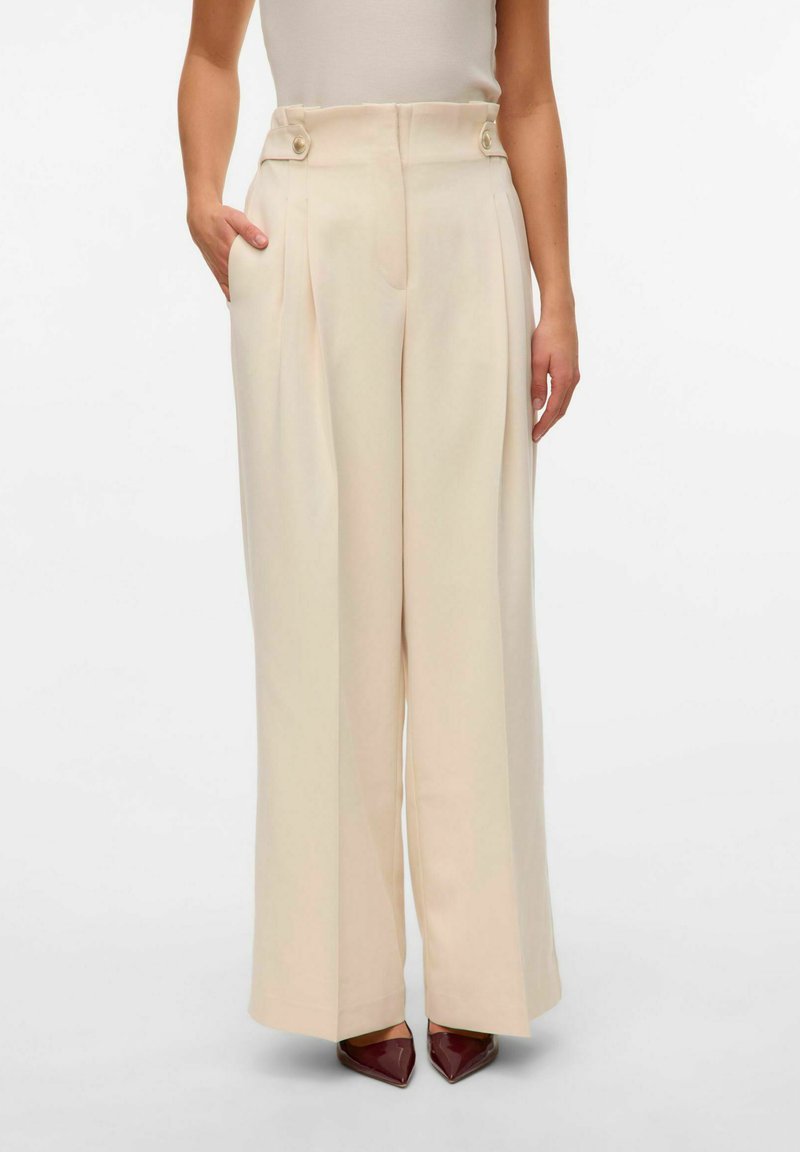 Pantalons larges en cream avec des plis à l'avant, des poches latérales et des accents à double bouton à la taille haute. Tissu lisse avec une texture douce.
