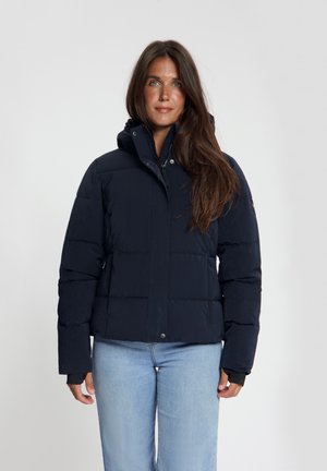 Vrouw met lang bruin haar, gekleed in een donkerblauwe donzen jas en lichtblauwe spijkerbroek, staand tegen een effen achtergrond.