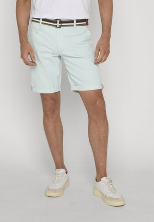 Mann trägt hellblaue Shorts mit aufgerolltem Saum, einen weißen Gürtel, weiße Turnschuhe und ein weißes T-Shirt, steht vor einem schlichten Hintergrund.