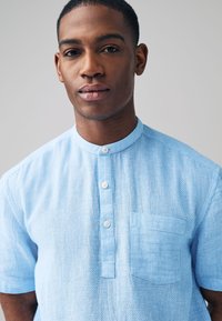 Lyseblå kortærmet skjorte med et tekstureret mønster. Har en opretstående krave, knapslå og en brystlomme. Stoffet virker blødt.