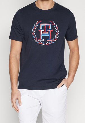 Mężczyzna noszący granatowy t-shirt z czerwono-biało-niebieskim logo "TH" otoczonym wieńcem laurowym, w połączeniu z białymi spodniami.