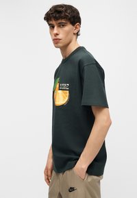 Νέος άνδρας με σκούρο πράσινο μπλουζάκι Nike με πορτοκαλί γραφικό και μπεζ παντελόνια Nike, στέκεται με το ένα χέρι στην τσέπη μπροστά σε απλό φόντο.