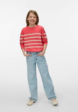 Smilende barn med lyst brunt hår iført en rød og beige stribet sweater, løse lyseblå jeans og beige slip-on sko, stående med hænderne i lommerne.