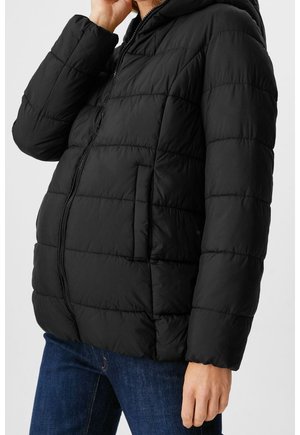 Person iført sort quiltet dunjakke med lynlås og forlommer, kombineret med mørkeblå jeans, hånd nær ansigtet.