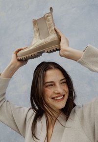 Bota Chelsea texturizada beige con un panel liso, suela gruesa y tira de agarre, sostenida contra un fondo azul claro.