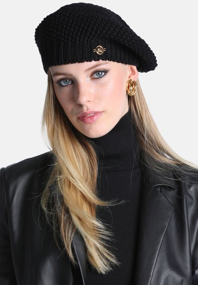 Femme aux longs cheveux blonds porte un béret en tricot noir, un col roulé noir, une veste en cuir noire et de grandes boucles d'oreilles dorées, regardant droit devant.