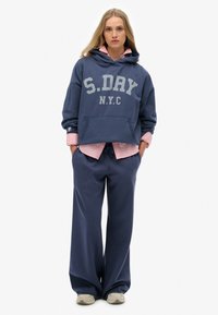 Sudadera con capucha de color azul marino con "S.DAY N.Y.C" impreso en gris, combinada con una camisa de botones rosa clara y pantalones deportivos en azul marino a juego.
