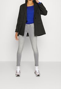 Zwarte dubbelgeknoopte blazer over een blauwe lange mouwen shirt, gecombineerd met lichtgrijze skinny jeans en witte sport sneakers.