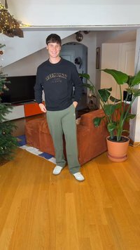 Sort sweatshirt med teksten "TOMMY JEANS", olivengrønne joggingbukser og hvide sneakers. Indendørs miljø med en brun sofa og plante.