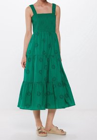 Kvinna klädd i en ärmlös grön midiklänning med ögonblicksblommönster och beige halkskor med dubbla spännen.