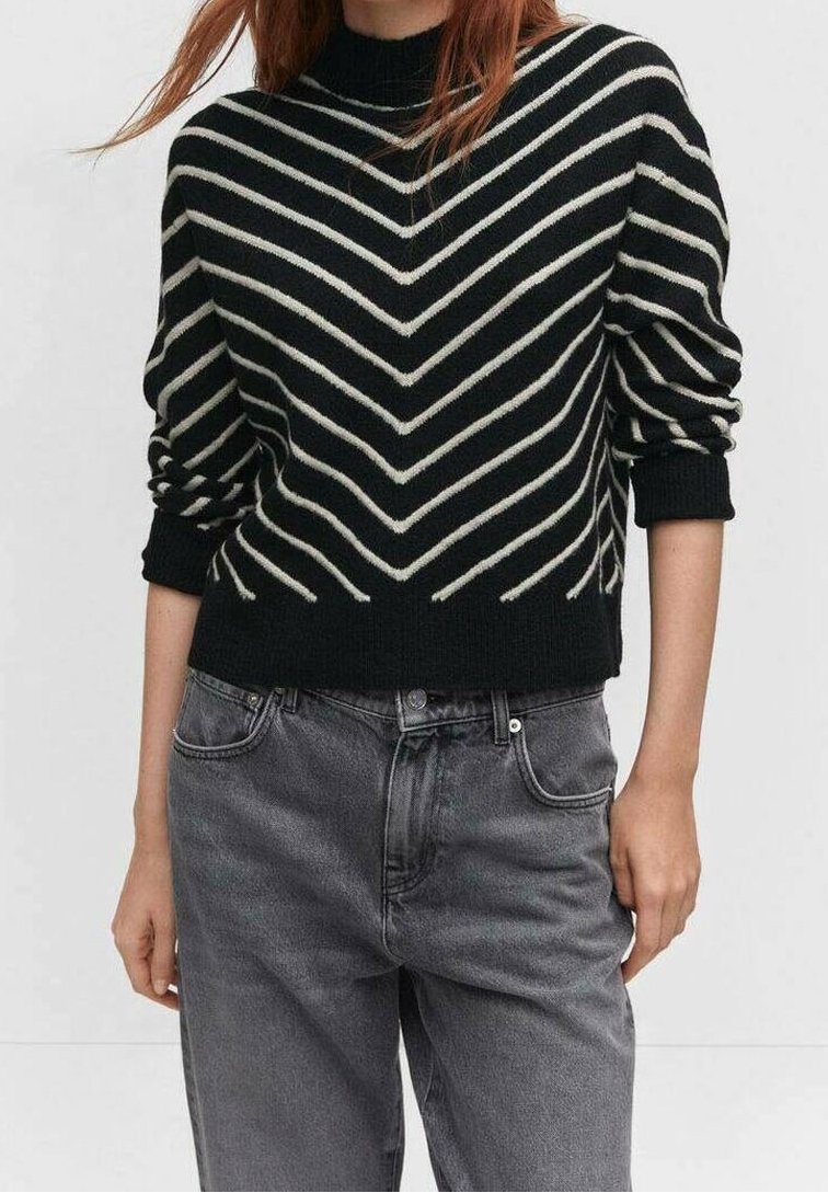 Femme portant un pull noir à rayures chevrons blanches et manches retroussées, associée à un jean gris délavé taille haute.