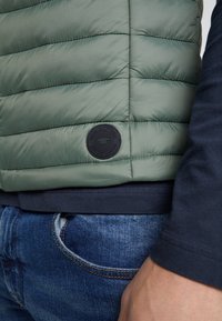 Fodrat grönt ytterplagg med horisontell quiltad design, texturerad tyg och ett runt svart logopatch på sidan. Bärs över en mörkblå skjorta.