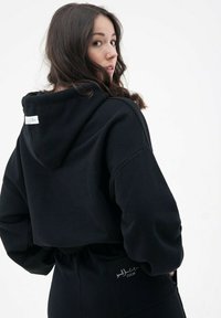 Czarny krótki hoodie wykonany z miękkiego materiału, z kapturem, luźnym krojem i metką na plecach. Towarzyszą mu pasujące spodnie.