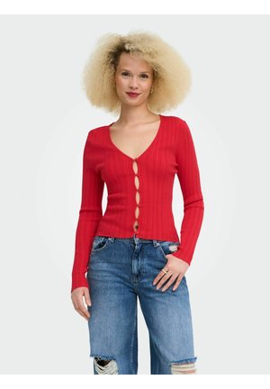 Femme aux cheveux blonds bouclés portant un cardigan rouge boutonné et un jean bleu déchiré, debout avec une main dans la poche.