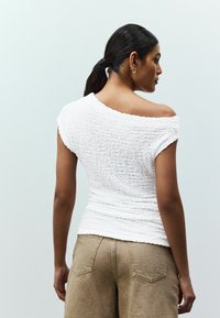 Bílý top s odhalenými rameny a texturovaným materiálem. Design obsahuje krátké rukávy a přiléhavý siluetu, doplněný béžovými kalhotami s vysokým pasem.