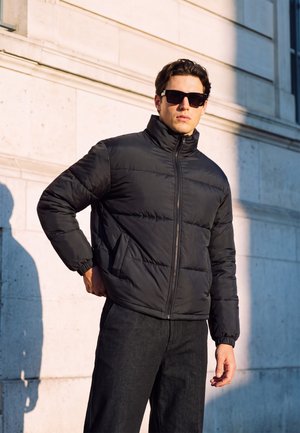 Jeune homme portant une doudoune noire et des lunettes de soleil, debout contre un mur en pierre éclairé par le soleil avec une ombre sur la moitié de son visage et de son corps.