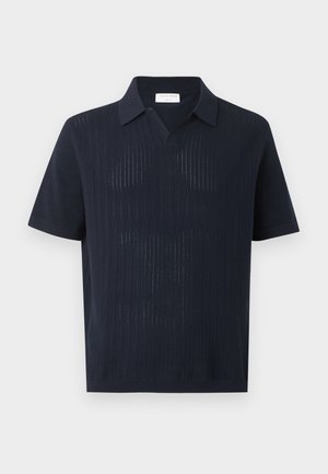 Homme portant une chemise noire à manches courtes avec col, pantalon vert olive et mocassins noirs, debout avec une main dans la poche.