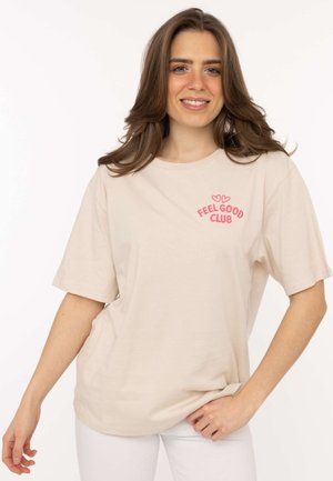 Frau mit schulterlangen braunen Haaren, die ein beiges "Feel Good Club" T-Shirt und weiße Hosen trägt, lächelt mit einer Hand auf der Hüfte.