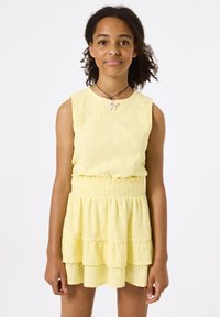 Robe sans manches jaune avec un motif floral texturé, taille smockée et ourlet en volants superposés. Portée avec un collier en coquillage.