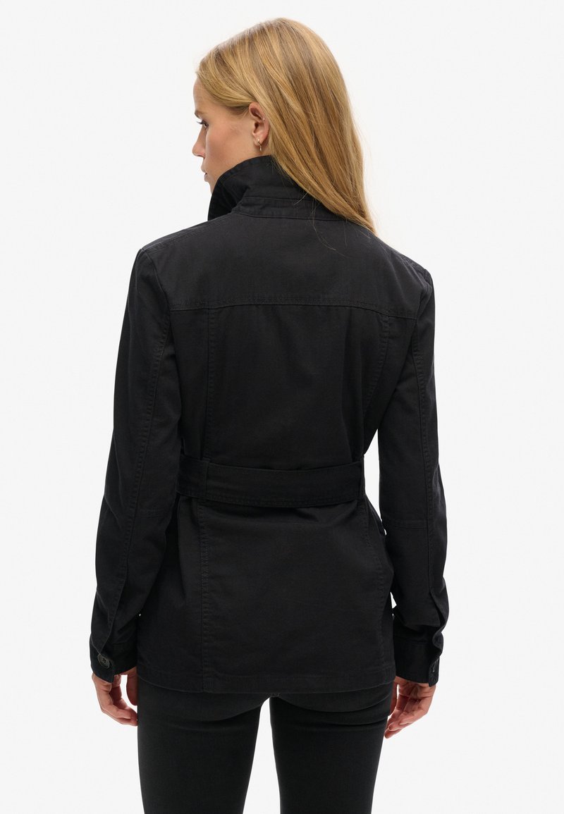 Chaqueta negra con cinturón, cuello alto y mangas largas. Presenta una textura suave y puños abotonados, mostrada desde atrás.