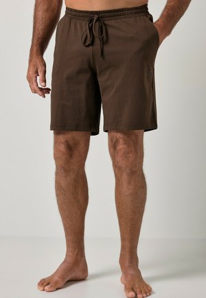 Mann trägt schlichte dunkelbraune Shorts bis zum Knie mit elastischem Bund und Kordelzug, steht barfuß vor neutralem Hintergrund.