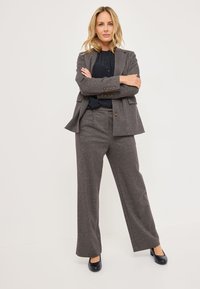 Completo grigio su misura realizzato in tessuto testurizzato, con giacca doppiopetto e pantaloni a gamba larga. Indossato con una blusa nera e tacchi blu navy.