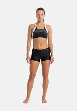 Donna in piedi a piedi nudi che indossa un reggiseno sportivo nero e bianco e pantaloncini neri aderenti contro uno sfondo bianco semplice.
