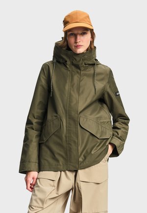 Donna con giacca con cappuccio verde oliva, pantaloni cargo beige e cappellino da baseball color fango, in piedi con una mano in tasca.