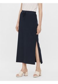 Femme portant une jupe midi bleu marine avec une fente latérale et une taille à cordon, associée à des sandales beige à talon bloc ouvertes sur les orteils et un haut blanc.