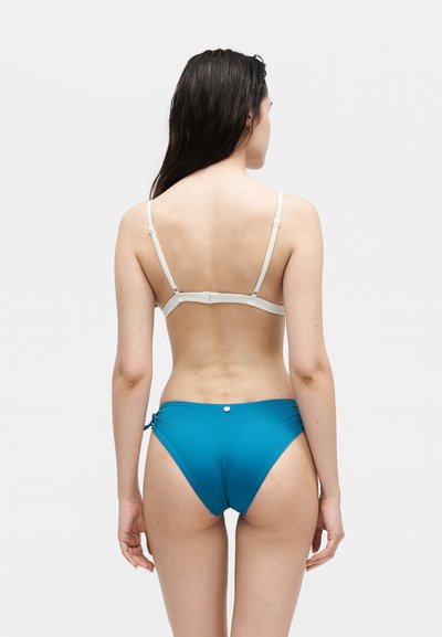 Γυναίκα που φαίνεται από πίσω, φοράει άσπρο τοπ μαγιό και μπλε κάτω μέρος μαγιό σε απλό ανοιχτό φόντο.