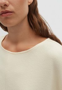 Frau mit langen braunen Haaren, die ein cremefarbenes, geripptes Strickoberteil mit rundem Ausschnitt trägt, dargestellt vom Kinn bis zur Schulter.