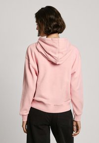 Felpa cropped rosa in cotone con cappuccio regolabile e polsini a coste, mostrata di spalle, abbinata a pantaloni scuri.