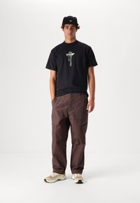 T-shirt nera in cotone con stampa grafica, pantaloni cargo marroni con tasche e sneaker beige con accenti neri. Il modello indossa un cappellino nero.