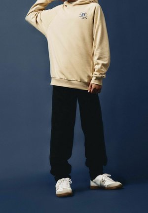 Sweat à capuche beige avec un logo sur le devant, pantalon de survêtement noir, et chaussures de sport blanches avec des accents gris. Tissu lisse, coupe décontractée.