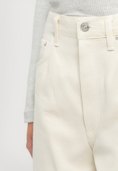 Detajlni posnetek osebe, ki nosi off-white visok pas jeans s kovinskimi gumbi in zakovicami, v kombinaciji s svetlo sivo rebrasto dolgo majico.