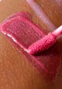 Glossy pink lip gloss being applied to brown skin with a doe-foot applicator, highlighting its shimmer and creamy texture.