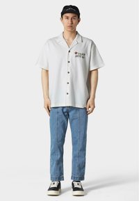 Camicia bianca a maniche corte con bottoni e ricami floreali, abbinata a jeans azzurri e scarpe da ginnastica nere. Cappellino nero regolabile in testa.