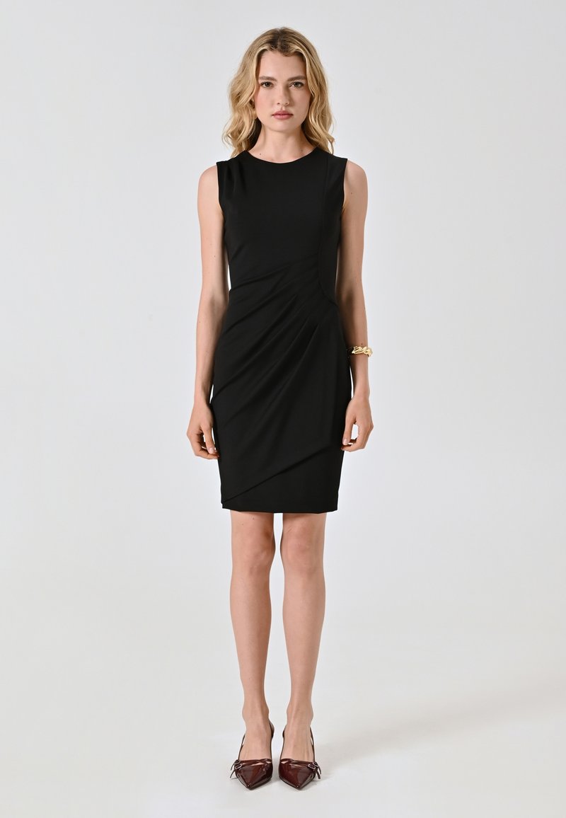 Schwarzes ärmelloses Kleid mit einer taillierten Passform und diagonalem Drapieren. Glattes Material und knielang, kombiniert mit burgunderfarbenen spitzen Absätzen.