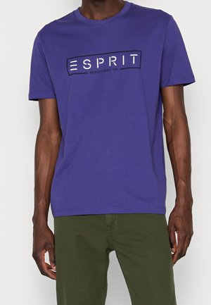 Mand iført en lilla T-shirt med korte ærmer og hvid "ESPRIT"-logo samt olivengrønne bukser, stående mod en ensfarvet baggrund.