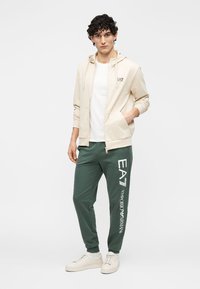 Beige hoodie met rits en logo, wit T-shirt, groene sweatpants met bedrukte branding en lichte sneakers. Glad stoftextuur.
