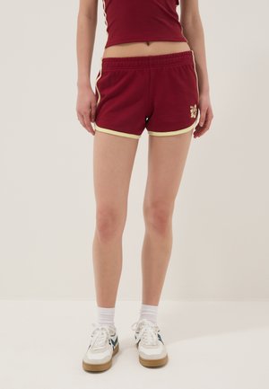 Persona vestida con pantalones cortos deportivos burdeos con ribete amarillo, una camiseta a juego, calcetines blancos y zapatillas blancas con detalles azules, de pie frente a un fondo liso.