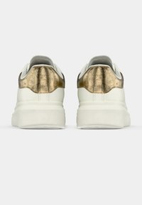 Baltas platforma sporta apavi ar metāliski zeltainiem papēža akcentiem, redzami no aizmugures uz vienkārša balta fona.