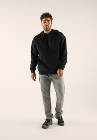 Sudadera negra con bolsillo tipo canguro y capucha con cordón, combinada con jeans grises y zapatillas deportivas negras. Diseño simple, textura suave.