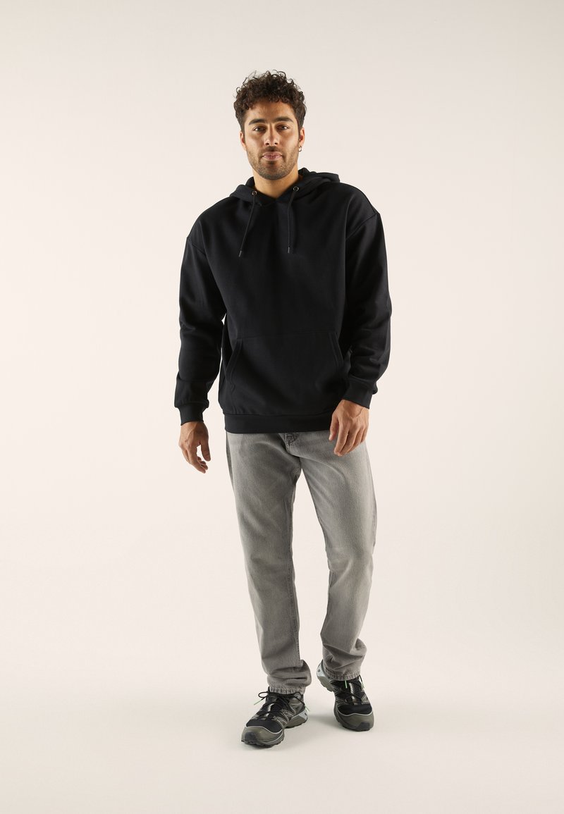 Sudadera negra con bolsillo tipo canguro y capucha con cordón, combinada con jeans grises y zapatillas deportivas negras. Diseño simple, textura suave.