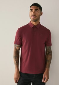 Polo shirt bordeaux a maniche corte, con chiusura a due bottoni e logo ricamato sul petto. Il tessuto è liscio con una sottile texture.