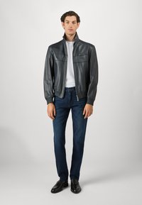 Άνδρας που στέκεται φορώντας μαύρο δερμάτινο μπουφάν, λευκό πουκάμισο, σκούρα μπλε τζιν και μαύρες τασέλες από παντόφλες σε ουδέτερο γκρι φόντο.