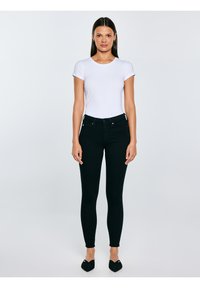 Kobieta stojąca na jednolitym tle, ubrana w białą koszulę z krótkim rękawem, czarne obcisłe jeansy oraz czarne spiczaste buty.