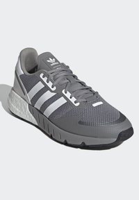 Šedá športová obuv s vrchnou časťou z pryžovej sieťoviny, troma bielymi pruhmi, polstrovaným golierom, reflexnými detailmi a textúrovanou gumovou podrážkou.