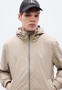 Μπεζ μπουφάν με κουκούλα και υφασμάτινη υφή, μπροστινό φερμουάρ και ελαστικές μανσέτες. Διαθέτει διακριτικό λογότυπο στο στήθος και πλαϊνές τσέπες.